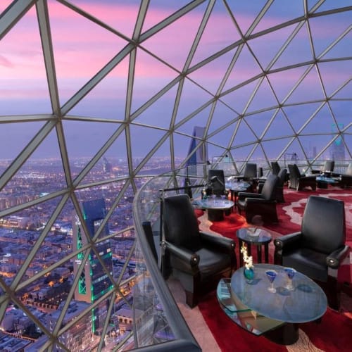 Luxushalle mit schwarzen Sesseln und Glastischen in einem Hochhaus mit geodätischer Glasskonstruktion, mit Panoramablick auf die Stadt bei Sonnenuntergang.
