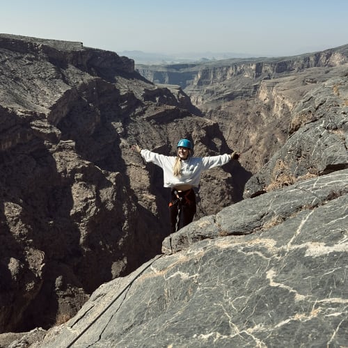 A person wearing a blue helmet and climbing gear standing with arms outstretched on a rocky cliff overlooking a deep canyon with rugged terrain under a clear sky.