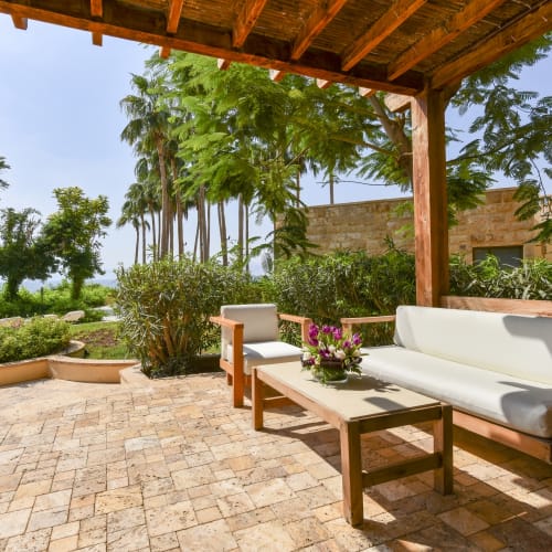Luxuriöse Terrasse im Freien mit Holzmöbeln, weißen Sitzkissen, Topfpflanzen und üppigem Grün unter einer Holpergola am Tag.