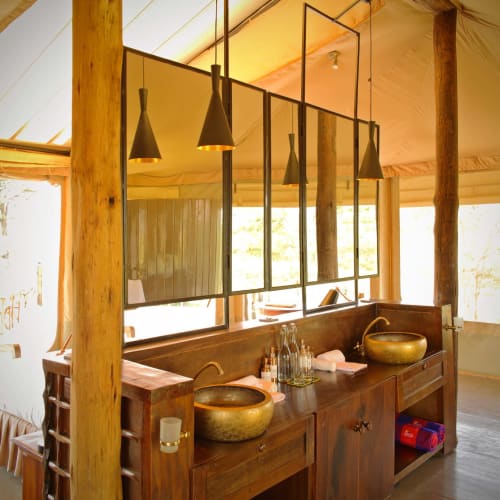 Elegantes Badezimmer mit zwei goldenen Waschbecken, hängenden Pendelleuchten und einer Holzwand im Zelt-ähnlichen Raum mit großen Fenstern.