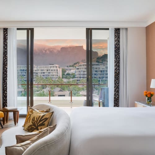 Ein modernes Hotelzimmer mit einem großen Bett, einem bequemen Sessel, einem kleinen Tisch, und einem Balkon mit Blick auf eine Stadt und Berge bei Sonnenuntergang.