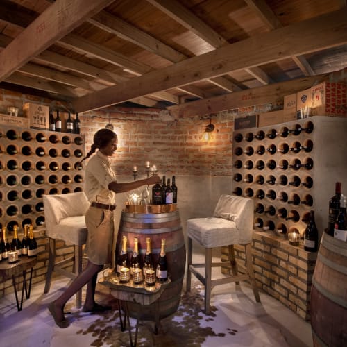 A woman is selecting a bottle of wine in a cozy wine cellar with brick walls and wooden beams.