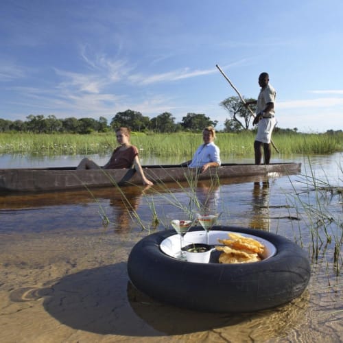 Drei Personen fahren in einem Mokoro-Boot auf einem ruhigen Fluss, eine Person steht und fischt. Im Vordergrund liegt ein Reifen mit Getränken und Snacks im Sand. Die Landschaft ist grün und üppig mit klarem blauen Himmel.