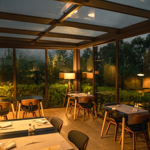 Cozy restaurant interior during the evening with large glass windows overlooking lush greenery, warm lighting, and neatly set tables.