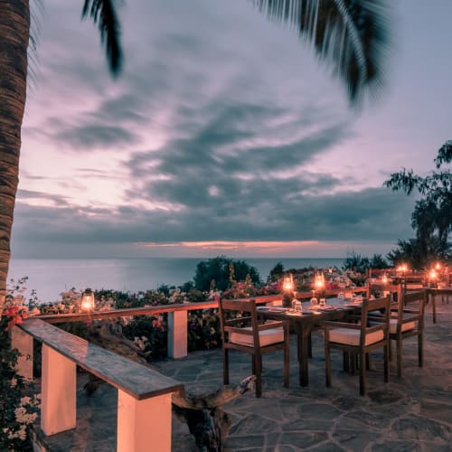 Picknicks im Freien auf einer Terrasse mit Tischen und Stühlen, mit Blick auf den Ozean bei Sonnenuntergang, in der Nähe von Palmen und einem Strohdach.