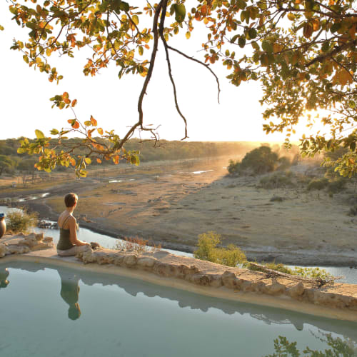 Eine Person sitzt friedlich am Rand eines natürlichen Beckens in Botswana bei Sonnenuntergang, umgeben von Bäumen und einer trockenen Landschaft.