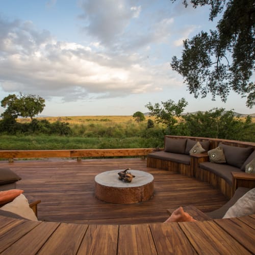 Ein runder Outdoor-Wohnbereich mit Holzsitzelementen, Kissen und einem Couchtisch auf einer Terrasse mit Blick auf die Savannenlandschaft.