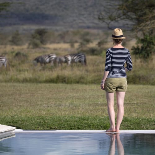 Eine Frau in gestreiftem Hemd und Shorts steht am Rand eines Swimmingpools und beobachtet Zebras auf einer Wiese in einer Naturreservat.