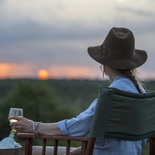 A woman wearing a hat and casual clothes sits in a chair holding a glass of wine, watching a sunset over the landscape.