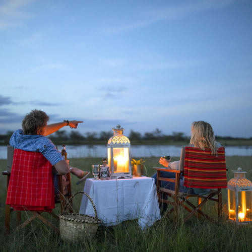Two older women are sitting outside at a table during twilight, with lanterns illuminating the scene while a man playfully points a toy gun at someone.