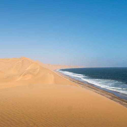 Vast desert dunes meet the ocean at Sandwich Harbour, Namibia, under a clear blue sky.