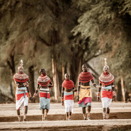 Six members of a local Samburu tribe walking in traditional attire on dirt ground with trees in the background.