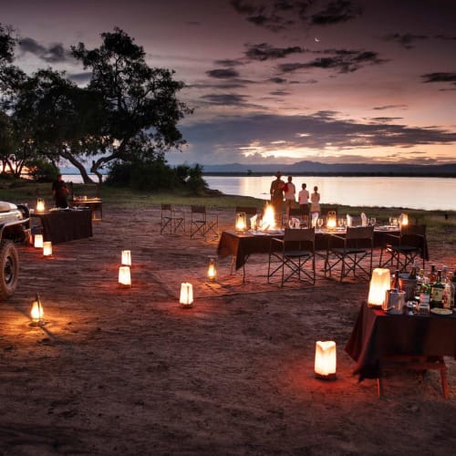 A romantic outdoor dinner setup on the riverbank at sunset, with tables, chairs, lanterns, and a bonfire, overlooking a tranquil lake.