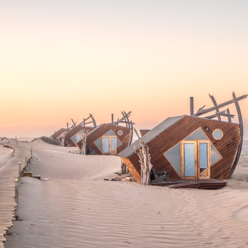 Abandoned boat structures on a sandy shoreline at sunset, resembling housed accommodations.