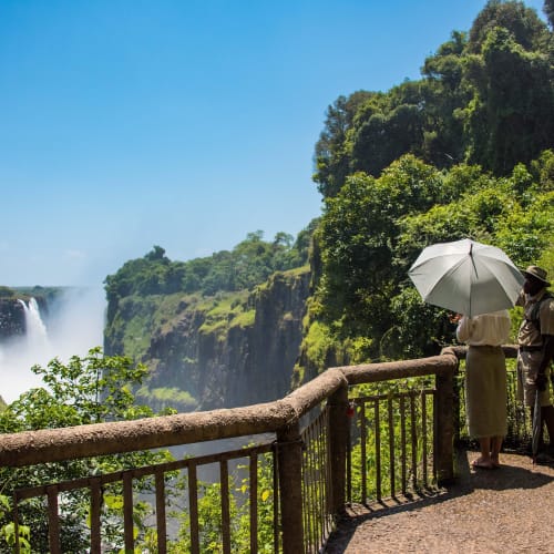 Visitors stand on a viewing platform overlooking Victoria Falls, surrounded by lush greenery under a bright blue sky.