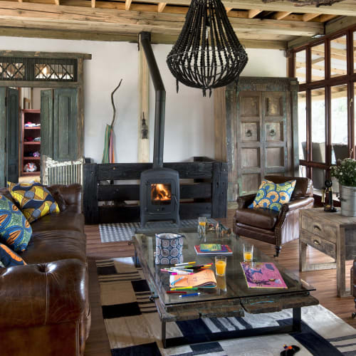 Cozy living room with a wood-burning stove, colorful cushions on leather sofas, and a rustic decor with wooden furniture and a woven chandelier.