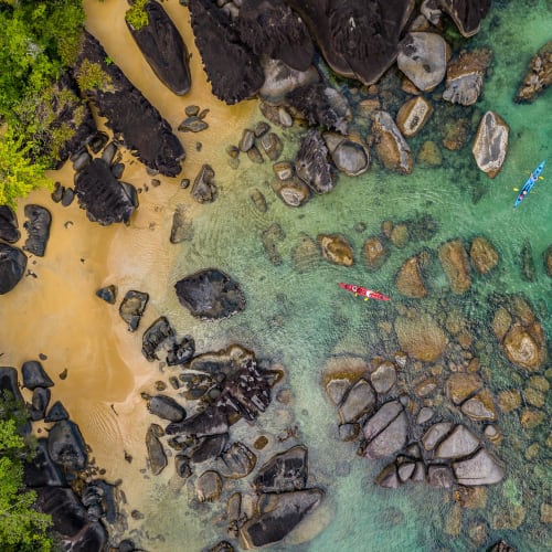 Colorful kayaks float on clear turquoise waters near a rocky and sandy shore surrounded by lush green trees.