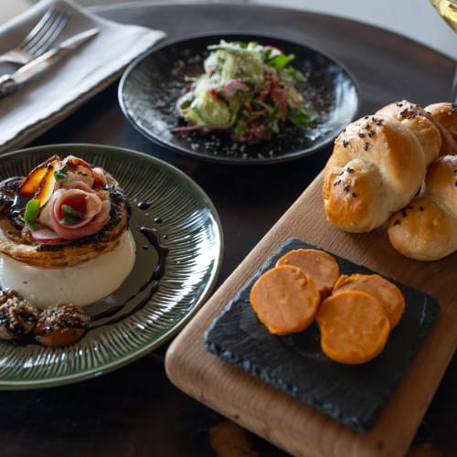 A gourmet dish with a poached egg topped with vegetables and sauce, accompanied by bread rolls, sweet potato slices, a fresh salad, and a glass of white wine on a dark table.