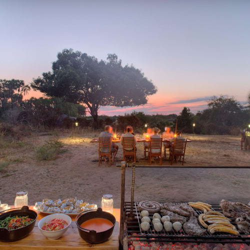 A group of people dining outdoors during sunset in a natural setting with a barbecue station in the foreground.