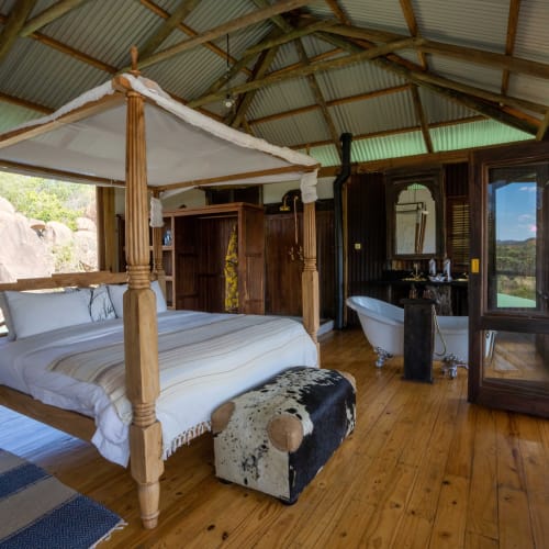 A cozy bedroom with a four-poster bed, wooden floors, and large windows overlooking a natural landscape.