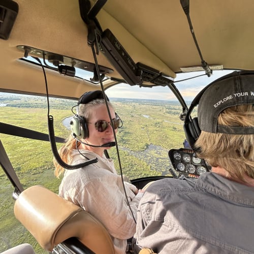 Two people wearing headsets inside a helicopter flying over a green, marshy landscape with scattered water patches.