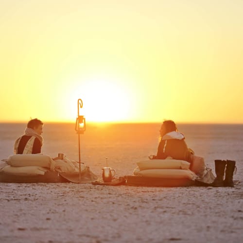 Zwei Personen sitzen auf Kissen, die einem Sonnenaufgang in einer flachen Landschaft zugewandt sind, mit Laterne und einigen Gegenständen in der Nähe.