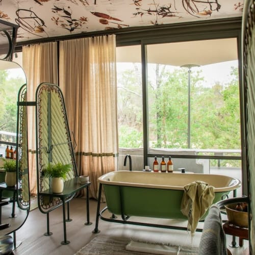 A spacious bathroom inside a tent with a green bathtub, large windows showing greenery outside, and various bottles and plants on the surfaces