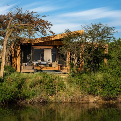 A modern wooden lodge situated on a riverbank amidst lush green trees in an outdoor setting.