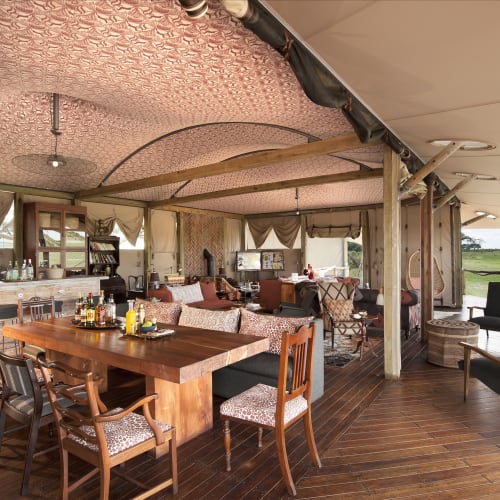 Freiluft-Hauptbereich eines Safari-Lodges mit Holzmöbeln, einem großen Esstisch und Blick auf grüne Felder.