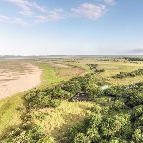 Aerial view of a lush green landscape with a small beachside lodge surrounded by dense trees and open fields in iSimangaliso, South Africa.