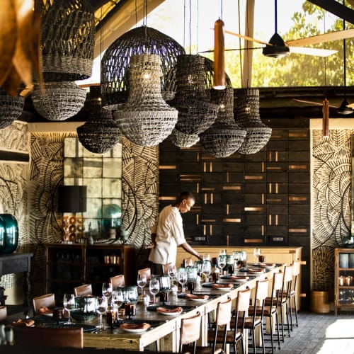 A sophisticated dining setup inside a well-decorated room with a person preparing food at a long table, featuring hanging woven lamps and patterned walls.
