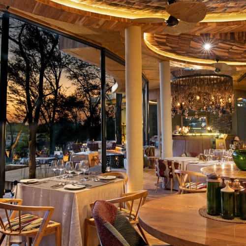 Elegant restaurant interior with sunset view, dining tables set with white tablecloths and glasses, stylish lighting fixtures, overlooking the natural landscape.