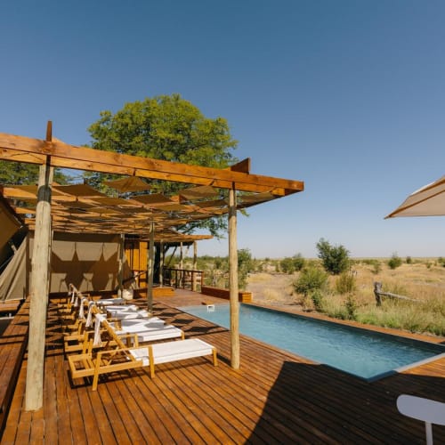 A luxurious outdoor swimming pool with wooden deck and lounge chairs surrounded by a natural landscape under a clear blue sky.