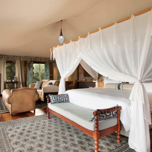 Luxurious bedroom with four-poster bed draped with white curtains, wooden furniture, and cozy seating area with armchairs and a sofa.