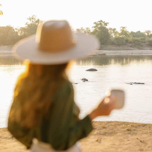 A woman wearing a large sun hat and a green jacket stands by a river, looking at her phone at sunset.