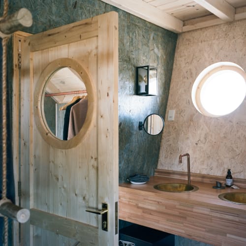 A rustic bathroom with a wooden partition featuring a circular mirror, two sinks, and a round window on a textured wall.