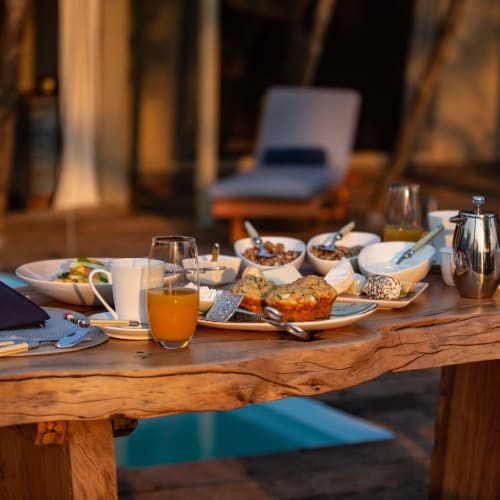 A rustic wooden table set for breakfast with various dishes, glasses, and utensils in an outdoor setting.