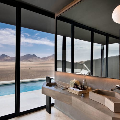Modern bathroom with dual sinks and large mirror overlooking a desert landscape through glass doors.