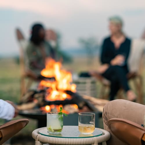 Menschen, die bei Sonnenuntergang Getränke um ein Lagerfeuer genießen, vor einer landschaftlichen Kulisse.