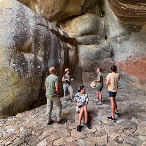 Eine Gruppe von sieben Personen erkundet eine felsige Höhle mit großen, glatten Felsen und einigen farbigen Malereien an den Wänden.