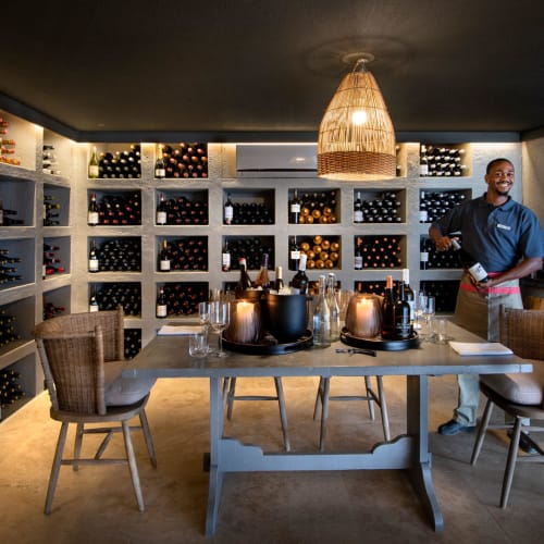 A wine cellar with shelves filled with wine bottles behind a table set for tasting, with a person standing nearby in a cozy room.