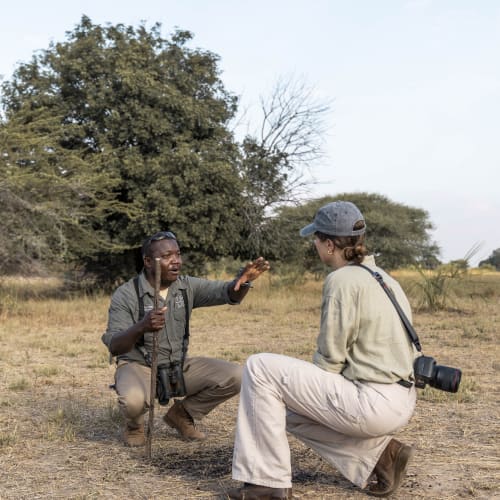 Ein Safari-Guide erklärt einer Touristin auf einer grasbewachsenen Fläche mit Bäumen im Hintergrund.