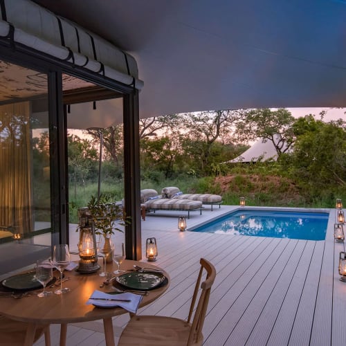 A cozy outdoor terrace with a wooden dining table set for dinner, lanterns lining the deck, and a swimming pool surrounded by nature at sunset.