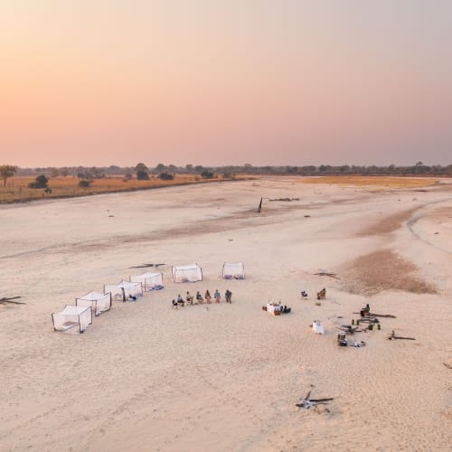 Eine Gruppe von Menschen sitzt in einem Halbkreis auf einem sandigen Gebiet bei Sonnenuntergang, mit mehreren Zelten und Ausrüstung im abgelegenen Gelände.