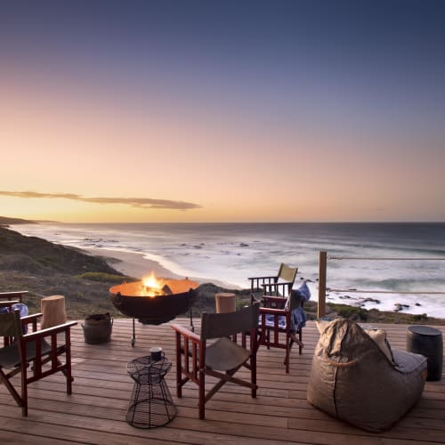 Gemütliche Sitzgelegenheiten auf einer Holzterrasse mit Blick auf das Meer bei Sonnenuntergang, umgeben von einem Feuerkorb und Strand, im Hintergrund das Wasser und der Himmel.