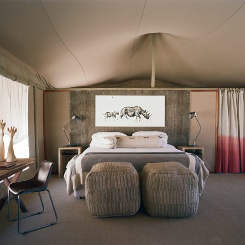 A cozy bedroom with a large bed flanked by two wall-mounted reading lamps, two ottomans at the foot of the bed, and a piece of animal artwork above the headboard, inside a tent-style interior.