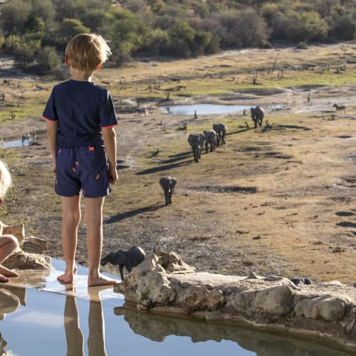 Zwei Kinder beobachten Elefanten an einer Wasserstelle in einer Savannen-Landschaft in Botswana.