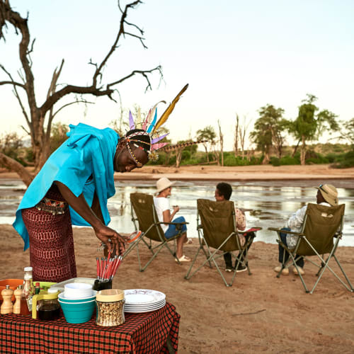A person dressed in traditional attire and a headdress preparing coffee on a table outdoors by a river, with three other people sitting on chairs in the background.