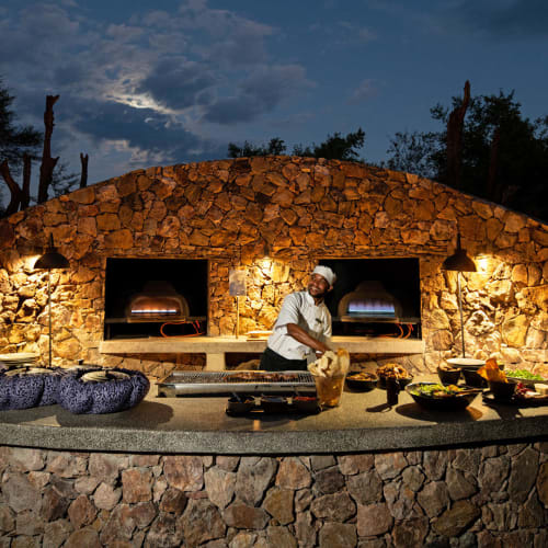 A chef preparing food at a stone outdoor kitchen with two brick ovens, illuminated at dusk, in a natural setting.