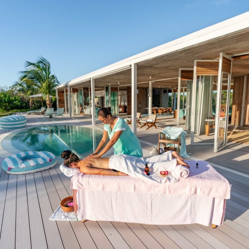 A woman receives a massage outdoors beside a pool at a resort with tropical trees and a modern wooden structure.